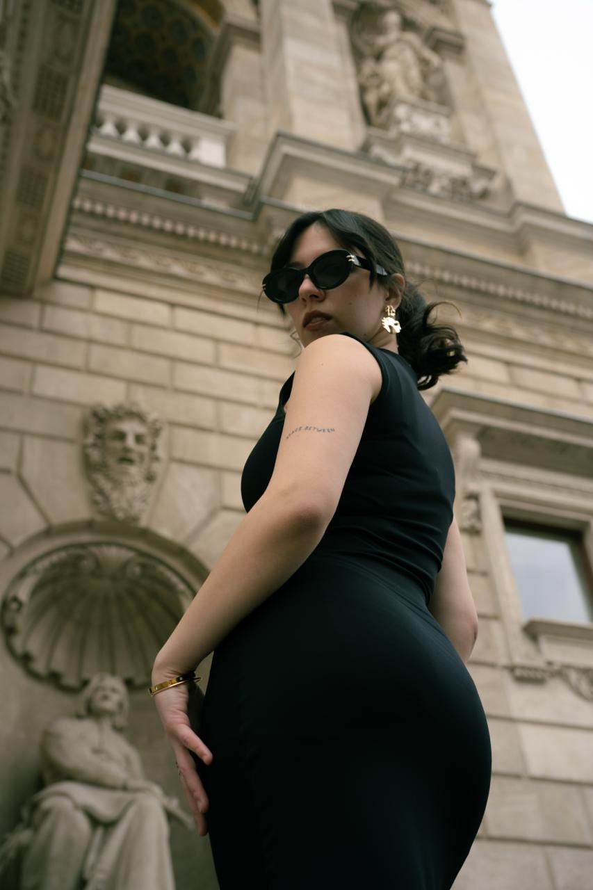A woman in sunglasses and a black dress, shot from below by a carved facade