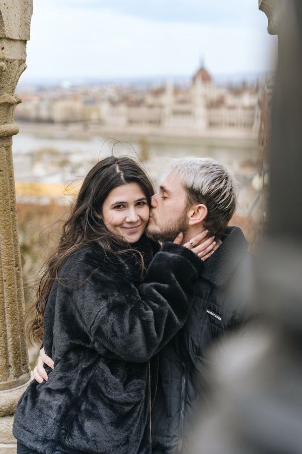 A man kissing a smiling woman on the cheek, the Parliament in the background