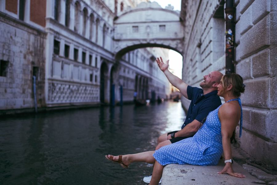 A couple sitting by a canal in Venice, the man pointing up at the Bridge of Sighs