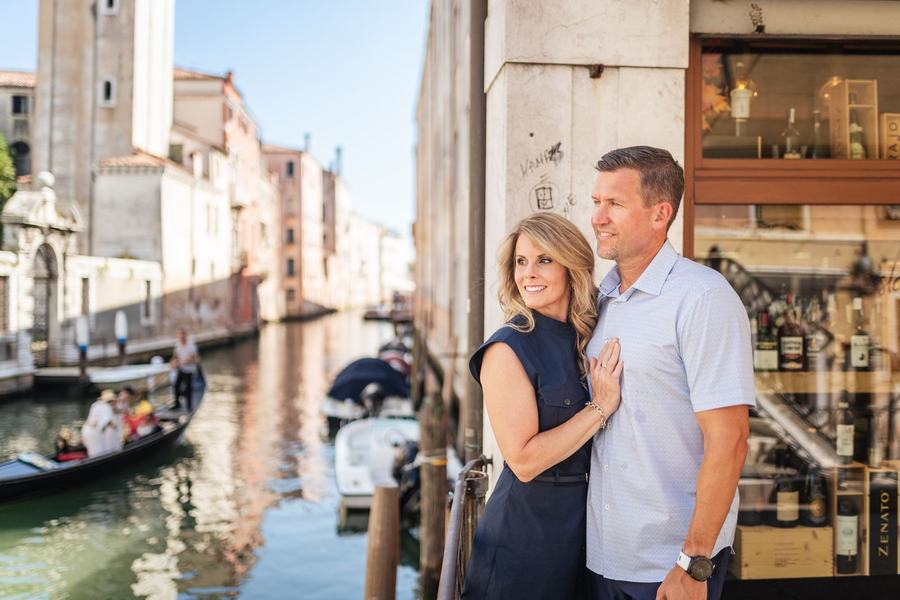 A couple standing by a canal next to a wine shop window, a gondola passing behind them