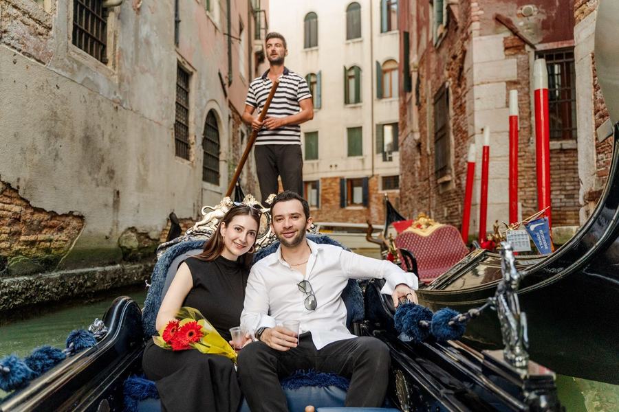A couple smiling on a gondola in a narrow canal, the gondolier behind them