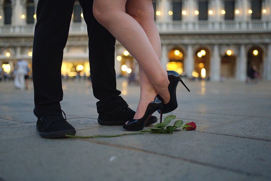 A woman in black heels lifting one foot as she leans into her partner in St Mark's Square at dusk