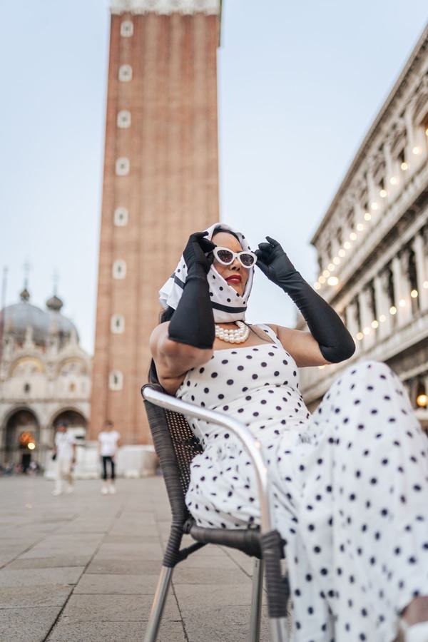 A woman in a polka-dot dress, headscarf, cat-eye sunglasses and long black gloves below the bell tower of St Mark's