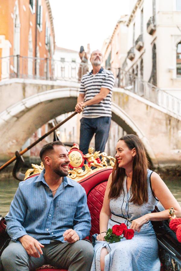 A couple laughing on a gondola under a small bridge, the woman holding red roses
