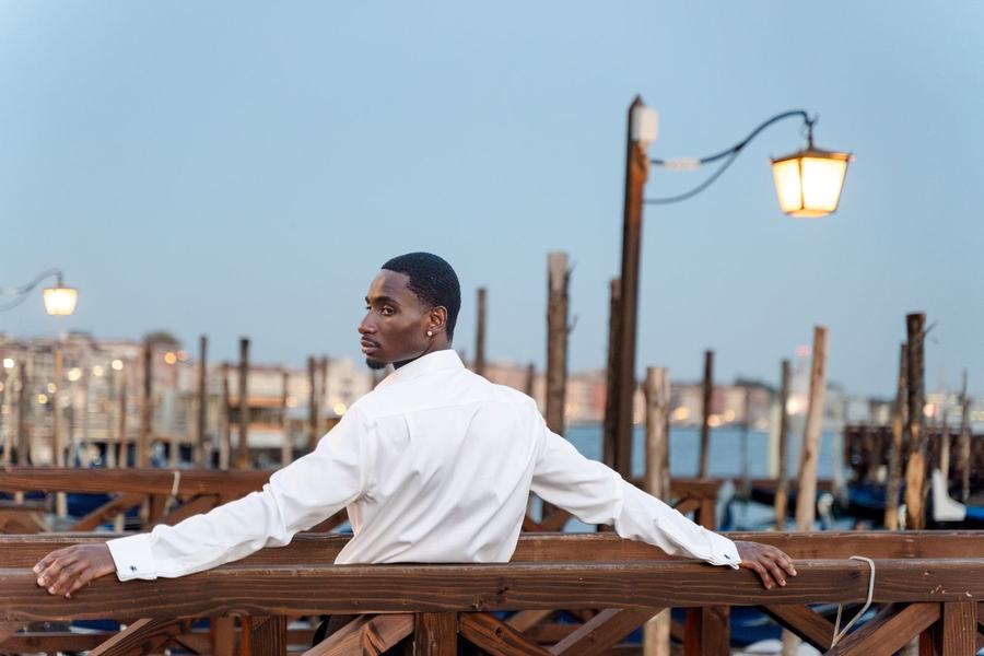 A man in a white shirt leaning on a pier railing at dusk, lanterns lit behind him